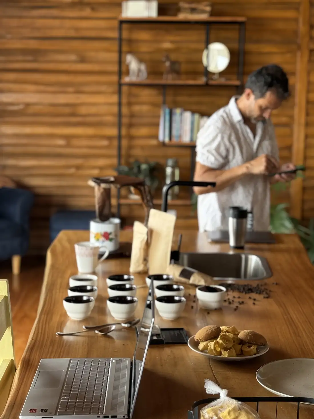 A taster standing at a long wooden table set with cupping bowls, noting impressions on a phone