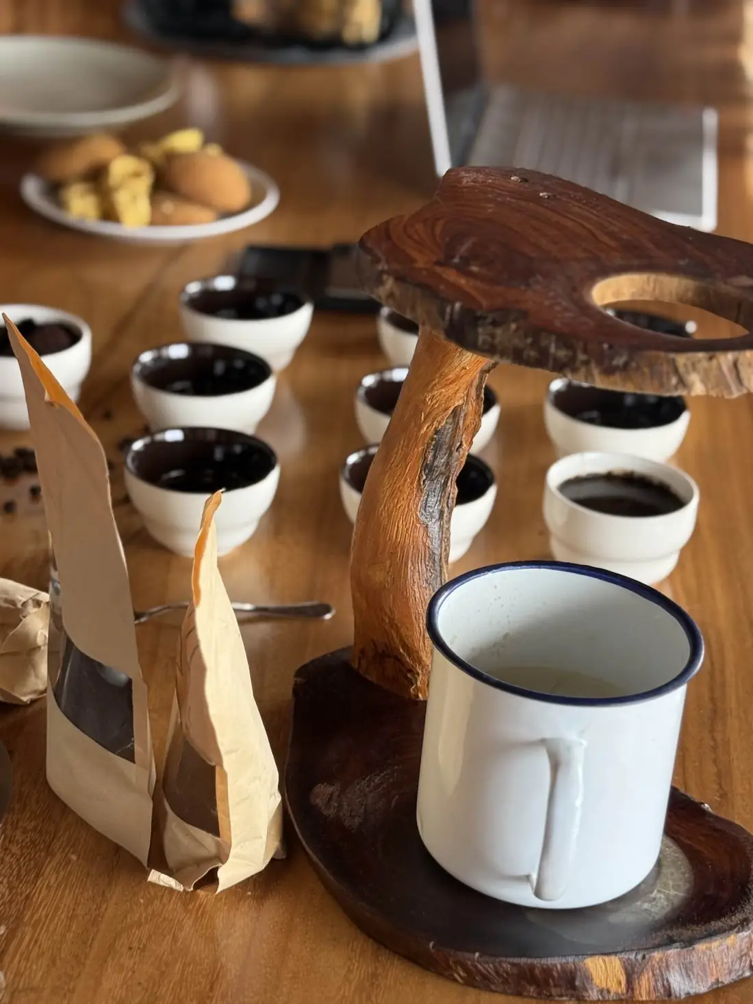 A pour-over on a wooden stand with an enamel mug and kraft coffee pouches beside cupping bowls